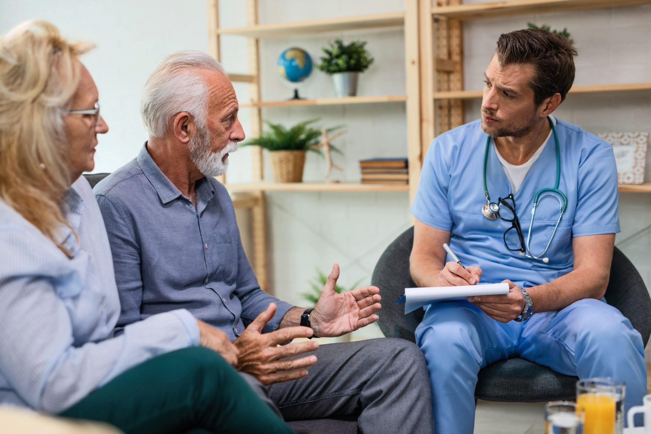 Ein älterer Mann und seine Frau erklären einem männlichen Arzt, der sie zu Hause besucht und sich Notizen macht, ihre gesundheitlichen Probleme.